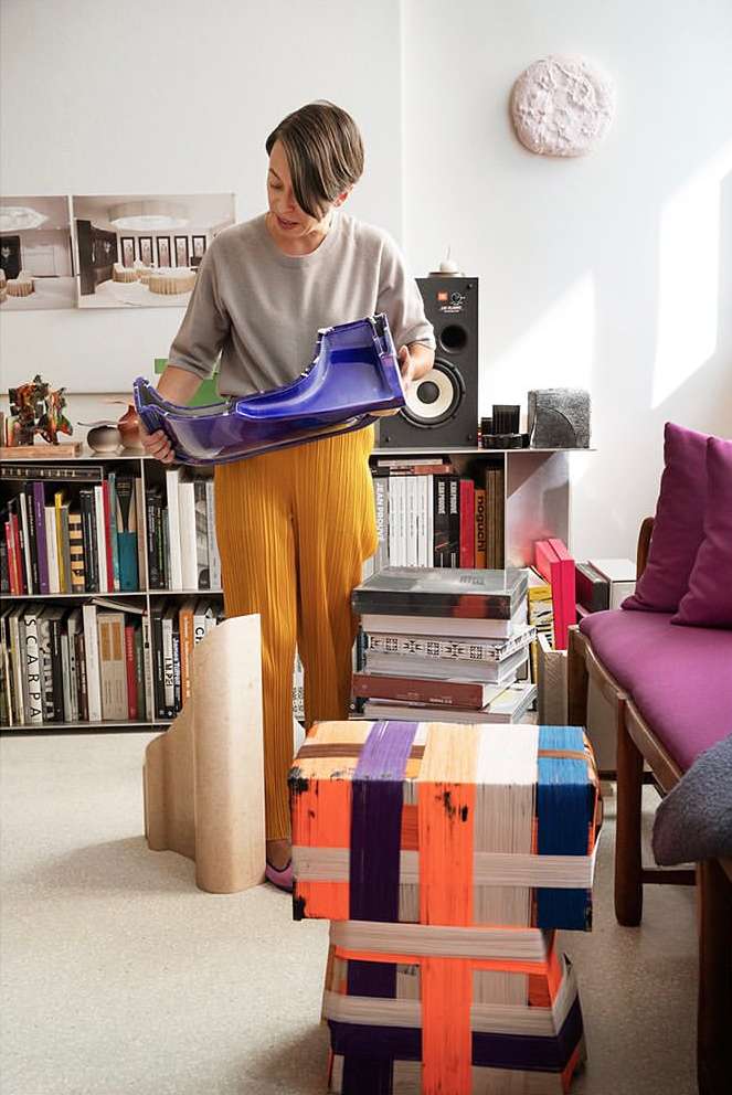 Woman in casual clothes holds a blue, molded plastic form in a bright room filled with books, speakers, and artful stacked stools.