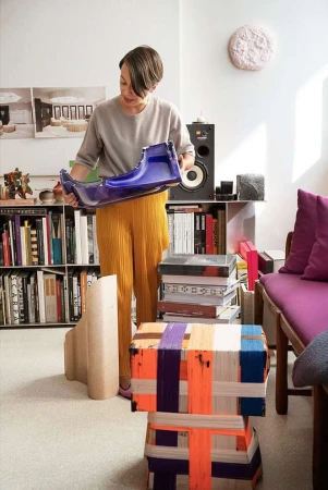 Woman in casual clothes holds a blue, molded plastic form in a bright room filled with books, speakers, and artful stacked stools.
