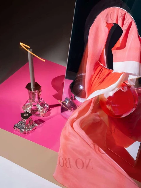 Still life photograph in studio featuring a candle in a glass holder, transparent balls, and coral-colored fabric on pink surface.
