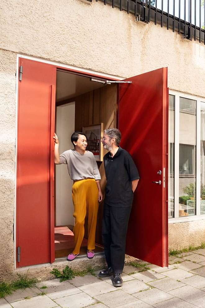 Two adults stand in a red doorway of a light beige building. An adult female smiles at an adult male. Natural light and a modern style.