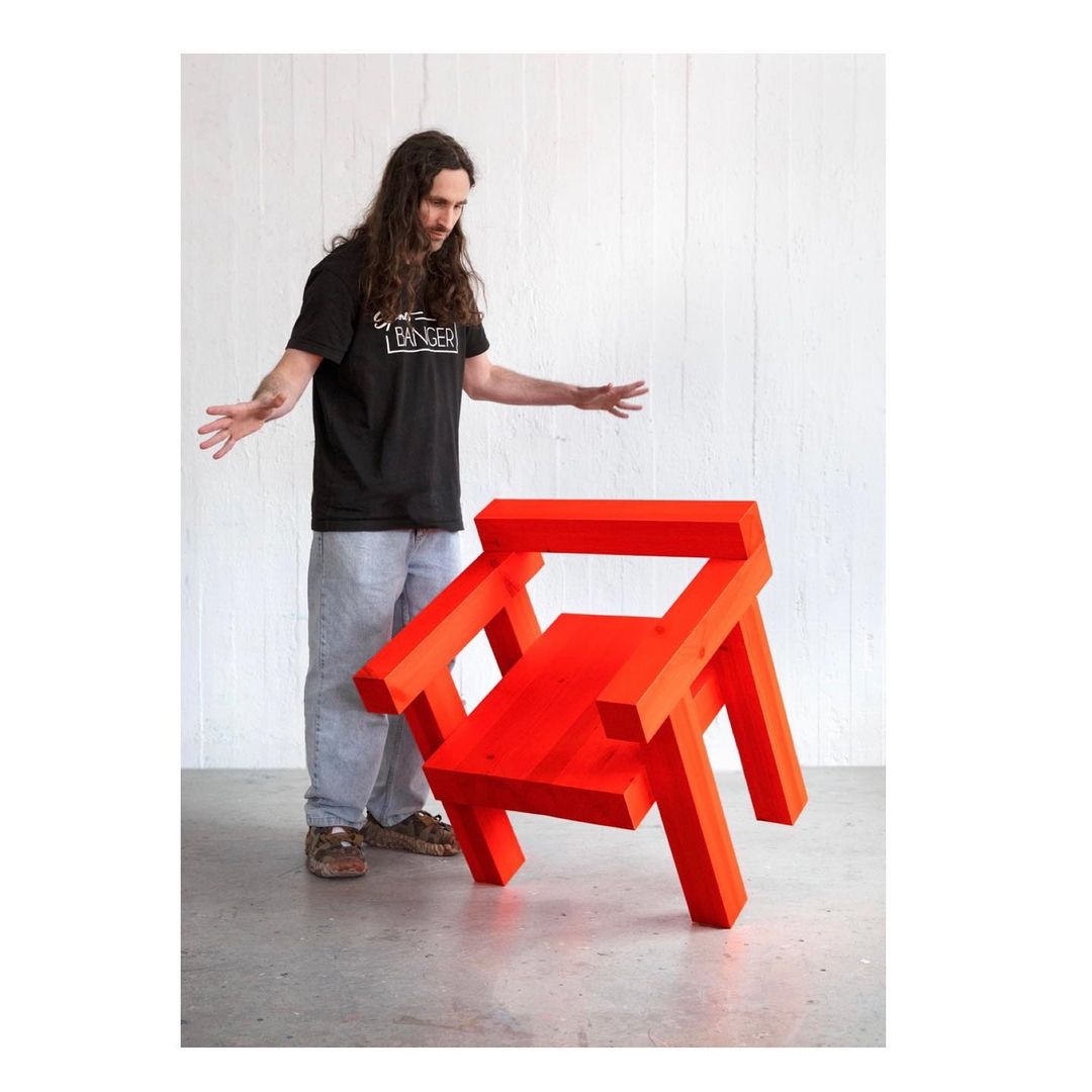Red angular deconstructed chair sits in front of a man in a black t-shirt and light blue jeans against a white wall indoors.