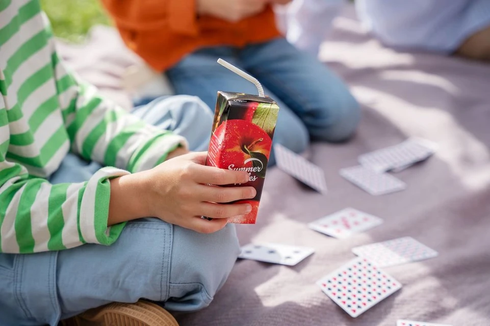 Child holding apple juice box outdoors with playing cards scattered on a blanket, casual setting, two children visible, daylight.