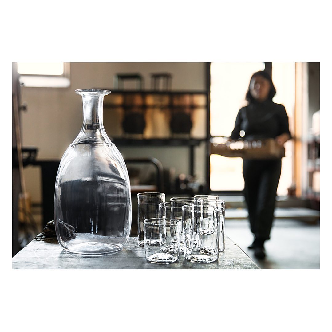 Clear glass carafe pitcher and group of glasses are set on a table with a blurred background featuring a person and window light.