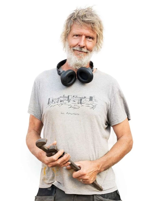 Adult male with a beard and grey t-shirt holding an adjustable wrench and wearing headphones. The background is plain white.