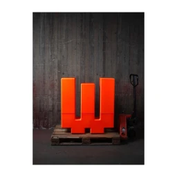 Bright orange sculptural block on a wooden pallet next to a pallet jack against a dark concrete wall