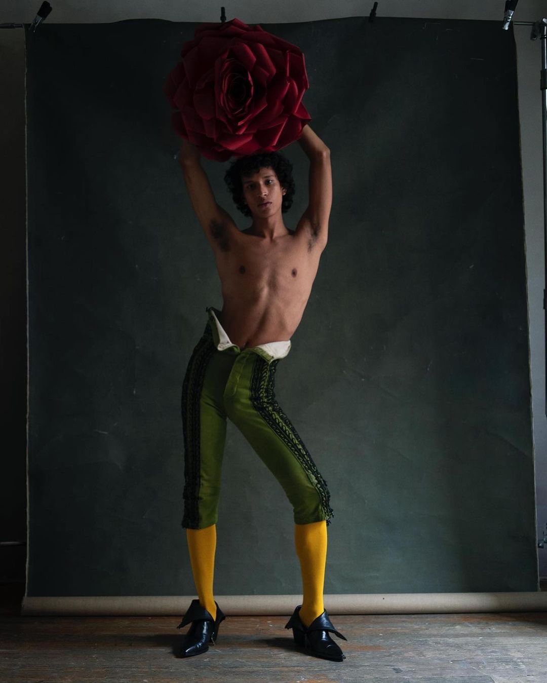 Man posing shirtless in cropped green pants, yellow socks, and black shoes, holding a large red flower in a studio setting.