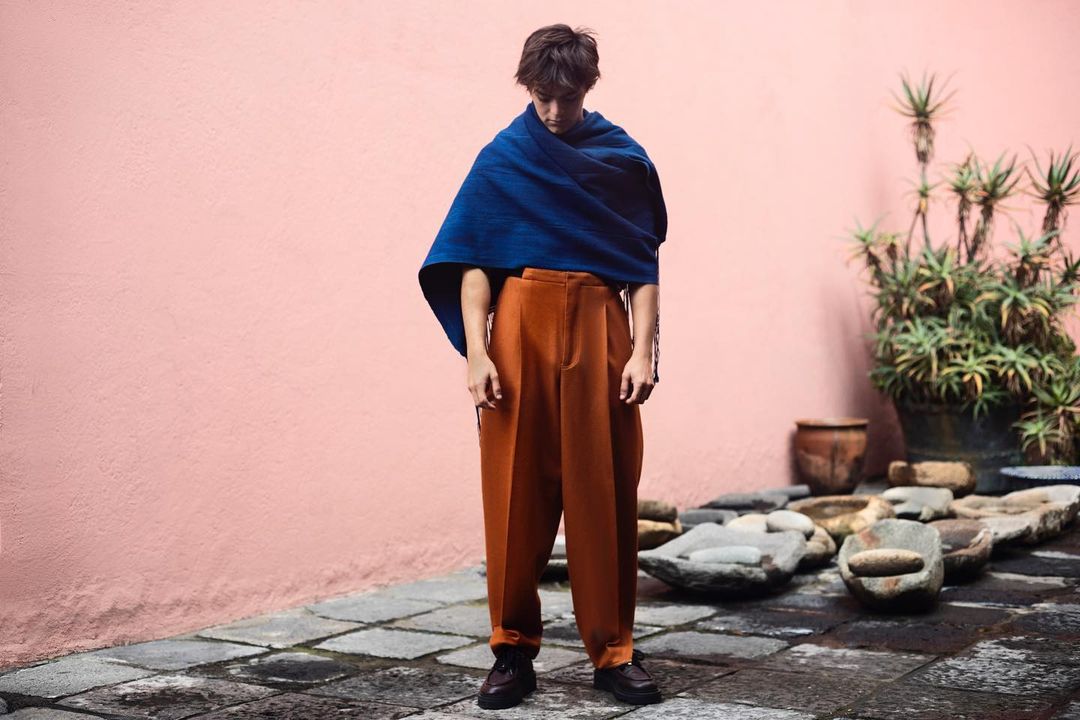 A young adult poses in burnt-orange pants and a blue shawl outdoors on a stone surface with a pink wall and green plants in the background.