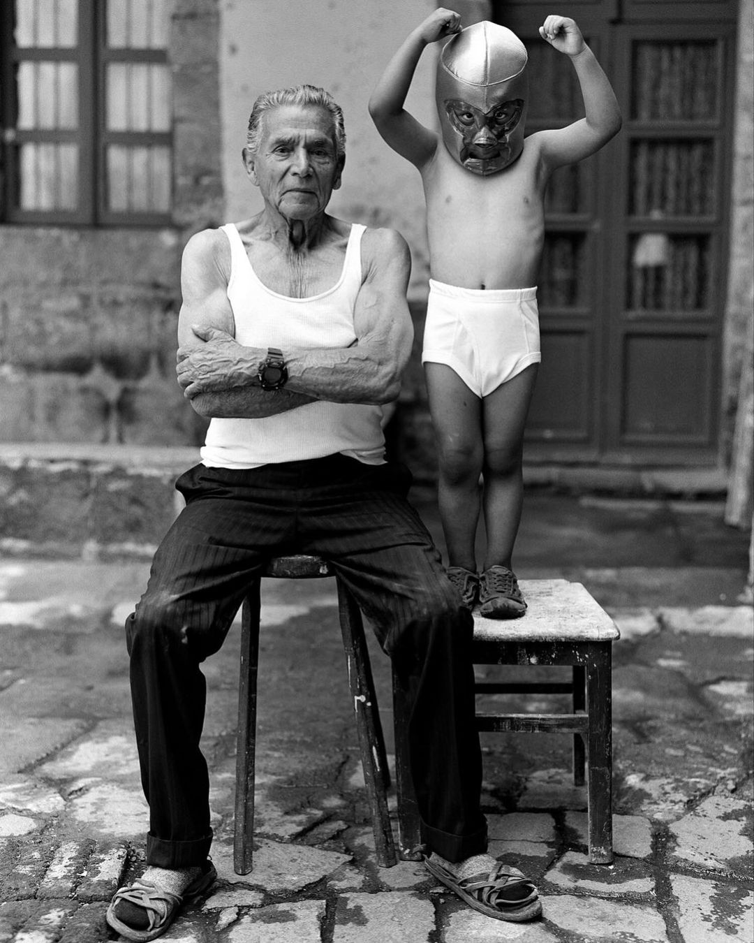 Black-and-white portrait of an elderly man seated on a stool next to a young child in a wrestling mask and briefs, evoking nostalgia.