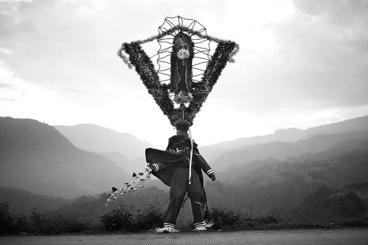 Black and white portrait of a costumed figure standing on a road with mountains in the distance, wearing a decorative headpiece.