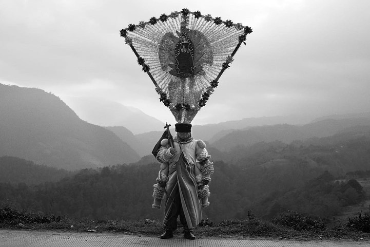 Black and white portrait of a person in a traditional costume holding a cross, standing against a mountain backdrop with overcast skies.