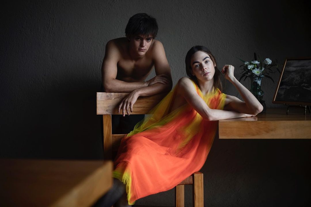 A fashion portrait featuring a topless man leaning on a chair and a woman seated at a desk in an artistic pose with natural lighting.
