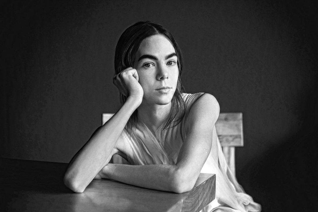 Monochrome portrait of a young adult with dark hair, wearing a light-colored dress, sitting at a table, with a dark background.