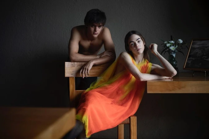 A shirtless man leaning on a wooden chair behind a woman seated in a bright orange and yellow dress.