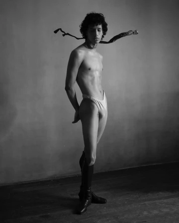 Monochrome portrait of a shirtless male model standing in tall black boots with a ribbon around his neck.