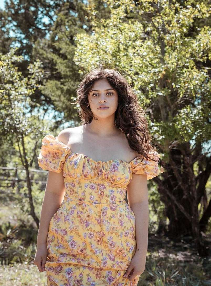 A woman stands outdoors in a yellow floral dress with puffy sleeves, posing in front of trees with green leaves and branches.