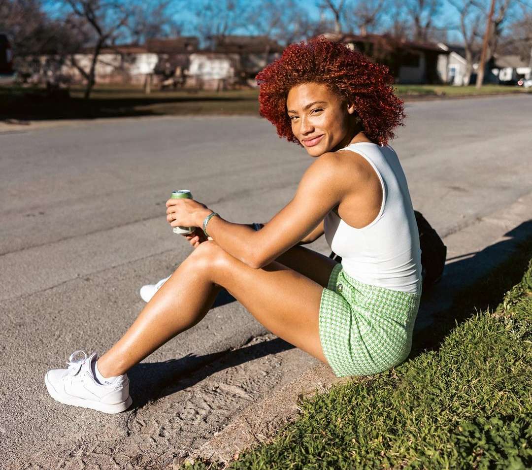 Red-haired woman in white top and green checkered skirt sits on a curb holding a drink outside with houses in the background.