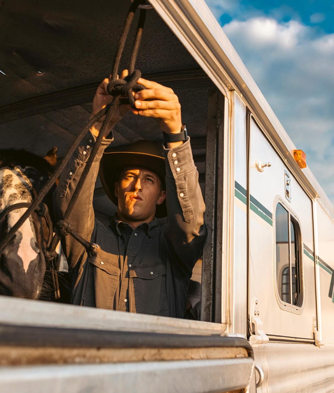 Cowboy securing rope inside horse trailer, preparing for an equestrian event with a cloudy sky background during golden hour.