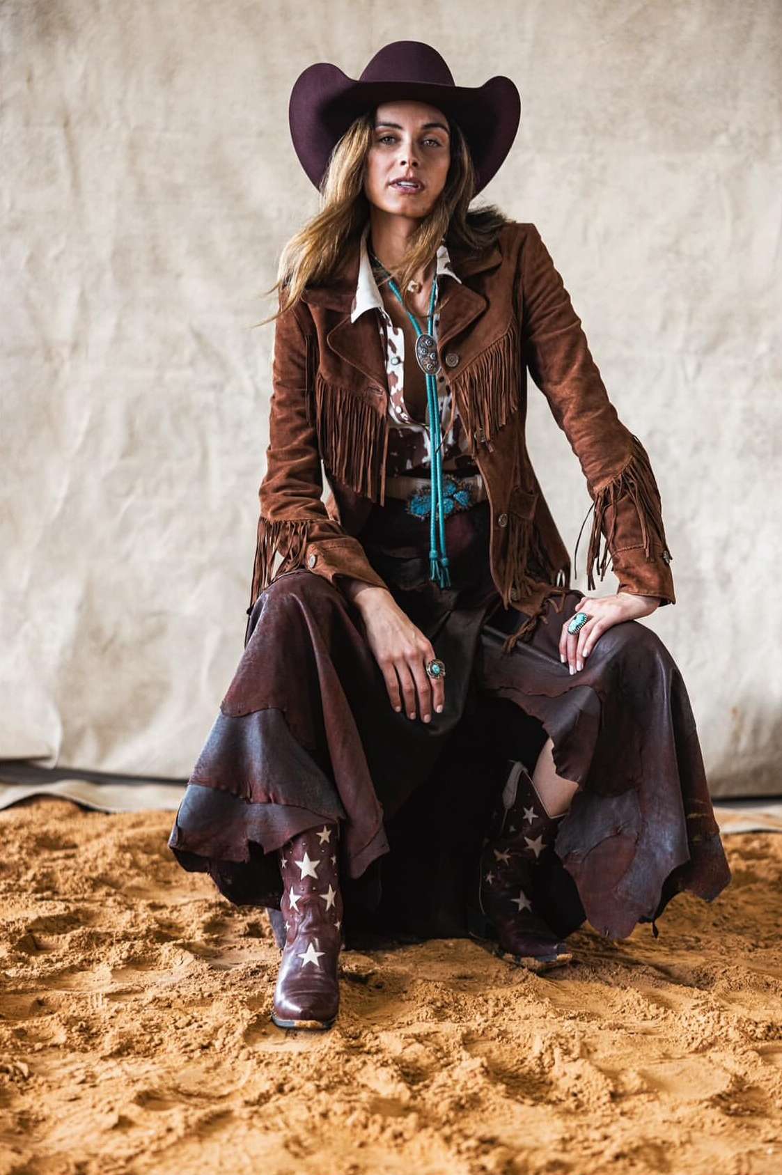 A woman in a cowboy hat, fringe jacket, and skirt sits on a sand-colored surface, looking directly at the camera in an outdoor setting.