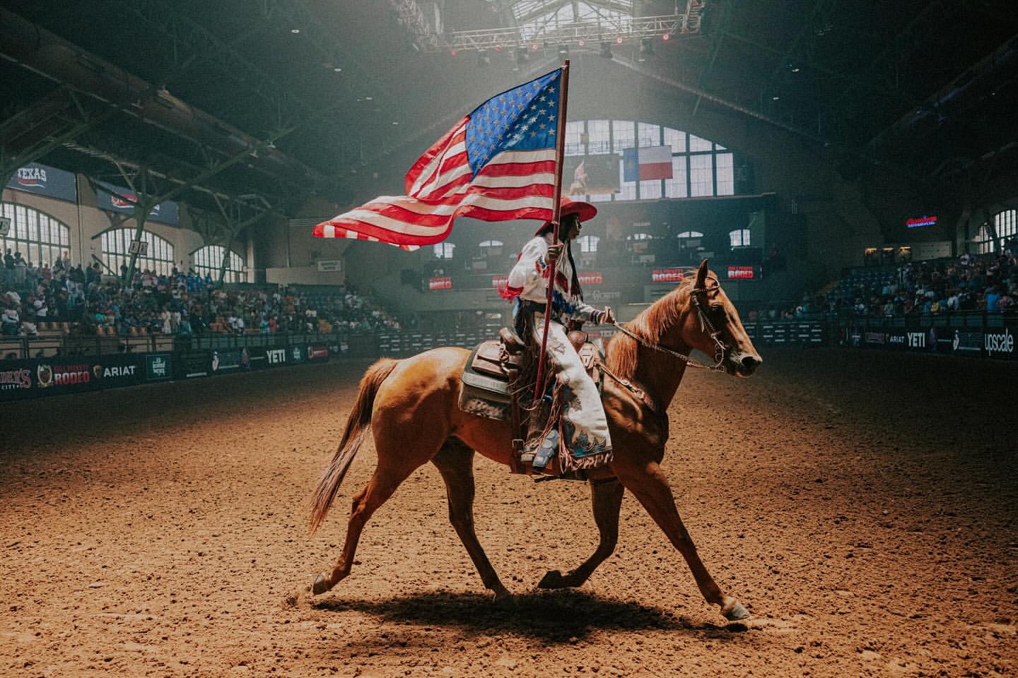 A horse and rider carrying an American flag in a rodeo arena with spectators, under bright spotlights, during a performance.