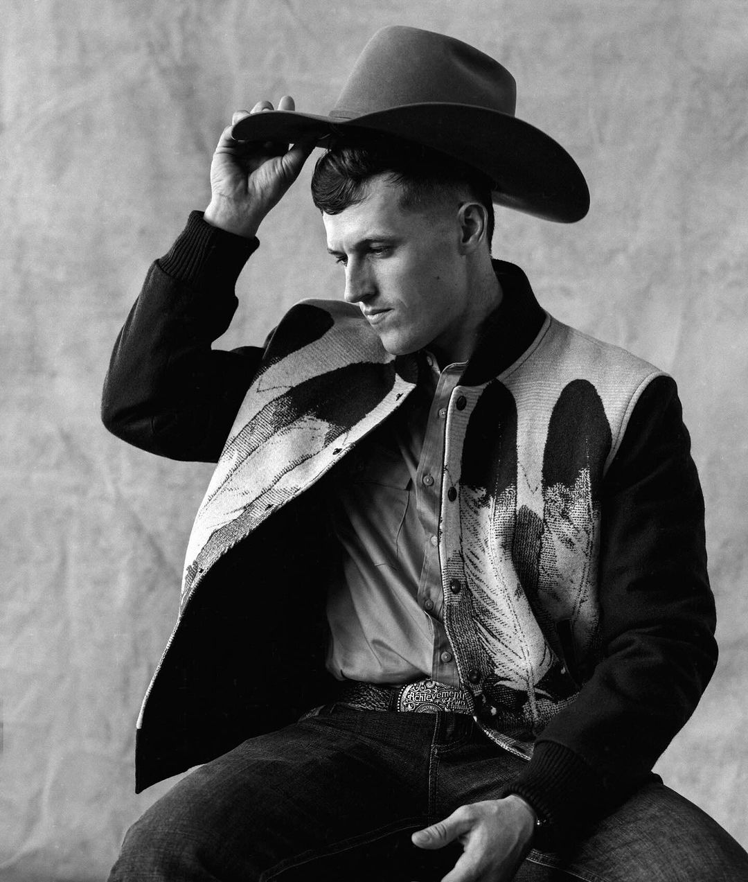 Portrait of a young man wearing a cowboy hat and patterned jacket, gazing downward, in a black and white studio shot.