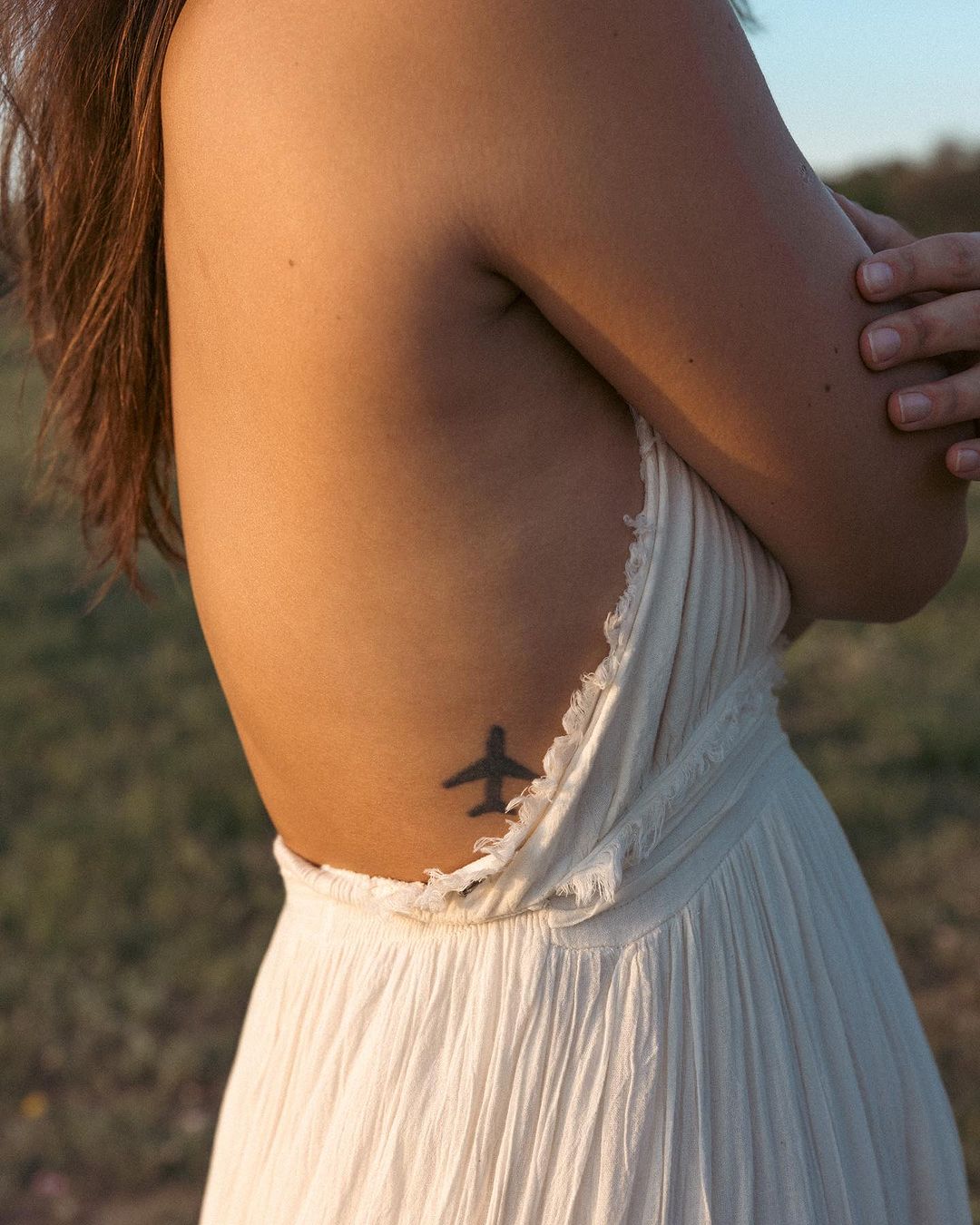 Woman in backless white dress with airplane tattoo on her ribcage standing in field. Soft natural light. Conceptual body art.