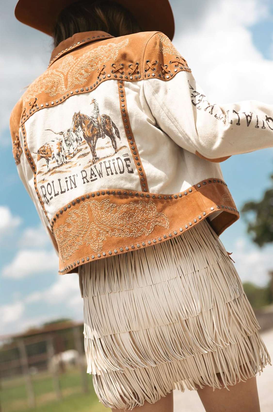 Woman wears a tan cowboy hat, tan and white jacket with western graphics, and fringed skirt, standing outside under a cloudy sky.