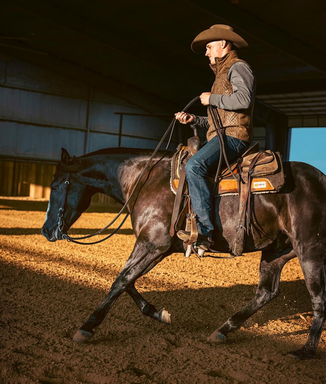 Adult male rides a dark horse in an indoor arena, wearing a cowboy hat and vest, with sand on the ground and some fencing in background.