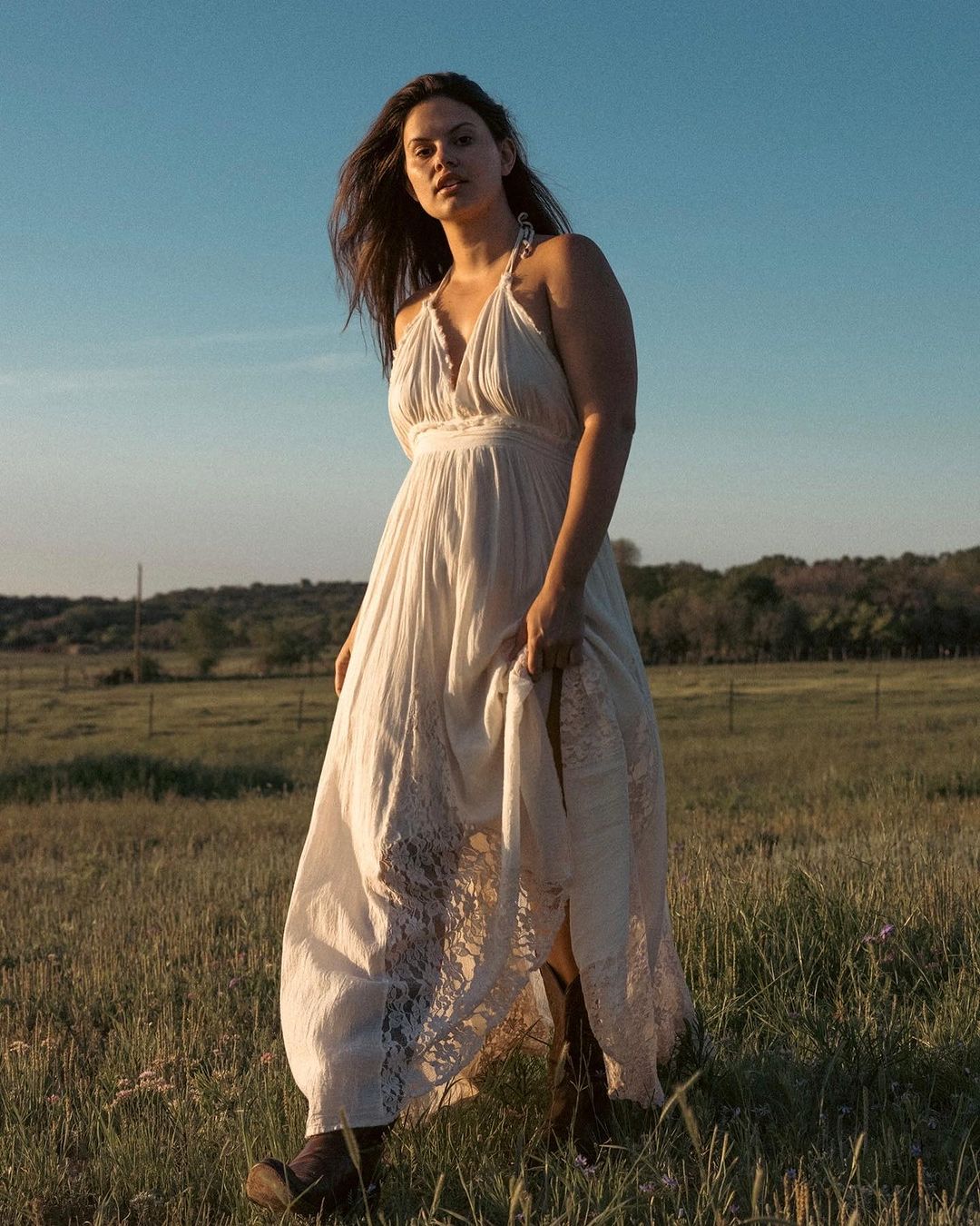 Woman in white lace dress and cowboy boots standing in a grassy field beneath a clear blue sky during golden hour.