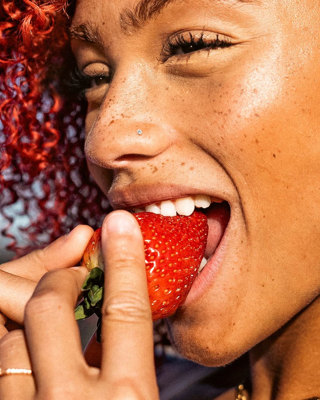 Close-up of a woman eating a strawberry with red curly hair, fair skin, freckles, and a nose piercing, focusing on skin and texture.