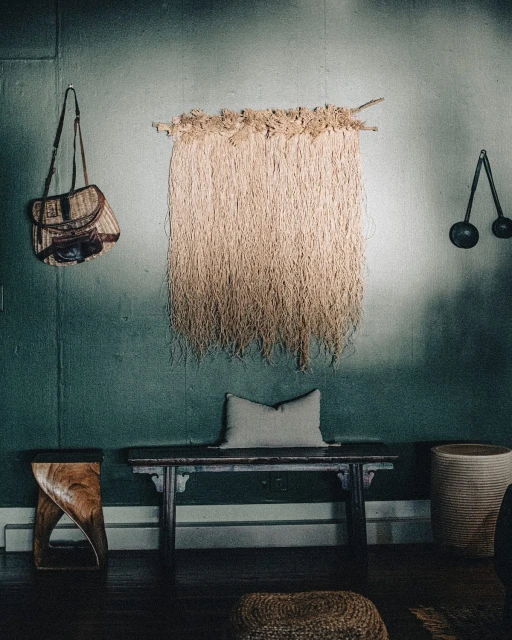 Interior wall decor: a woven raffia wall hanging, vintage purse, black iron sconces, wooden bench and stool against dark teal plaster walls.