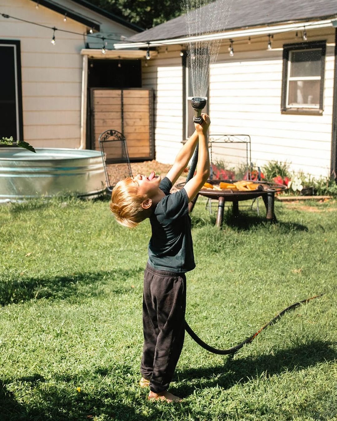 A blonde-haired child plays in the sunlit backyard, raising a garden hose overhead as water sprays in the air, laughing. House background.