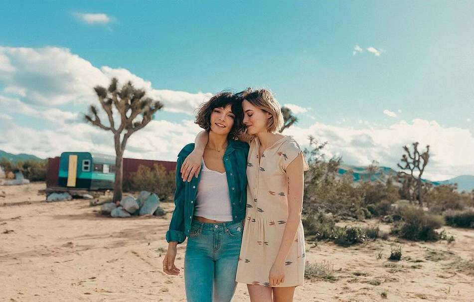 Two adult women embrace in a sun-drenched desert landscape with sparse vegetation under a bright blue sky filled with scattered clouds.