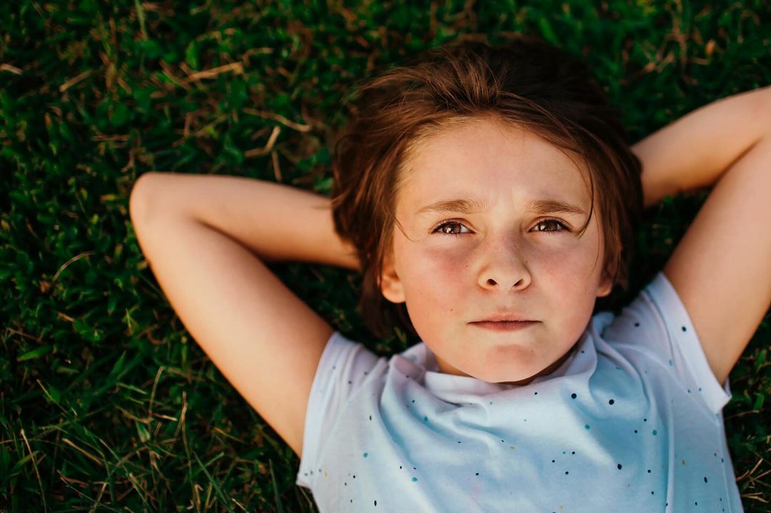 Young child lies in vibrant green grass outdoors with arms behind their head. Close up portrait, sunny lighting, and cheerful mood.