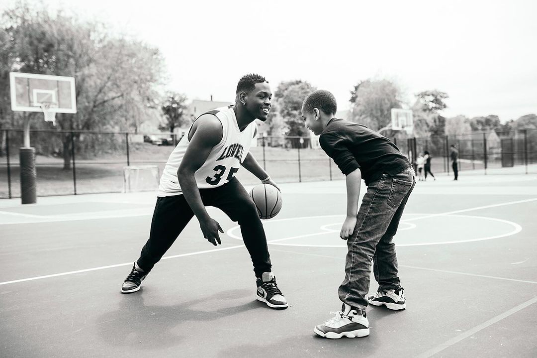Two African American men playing basketball on an outdoor court. The older man dribbles as the younger man defends in monochrome.