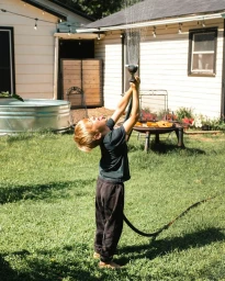 Young child holding a garden hose overhead with water spraying upward in a grassy backyard.