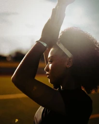 Profile portrait of a woman with curly hair raising her arms against a backlit sky by Sara Forrest