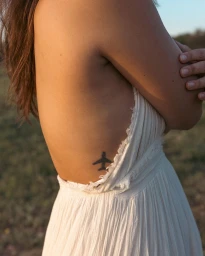 Close-up profile of a woman in a white pleated dress with an airplane tattoo on her torso, photographed by Sara Forrest.
