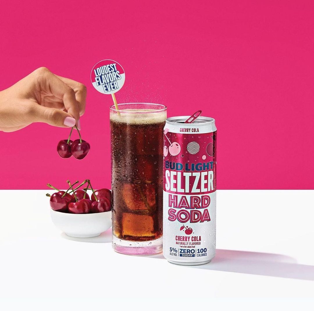 Bud Light Seltzer Hard Soda, cherry cola flavor, stands with glass of iced drink and cherries, set against a pink background.