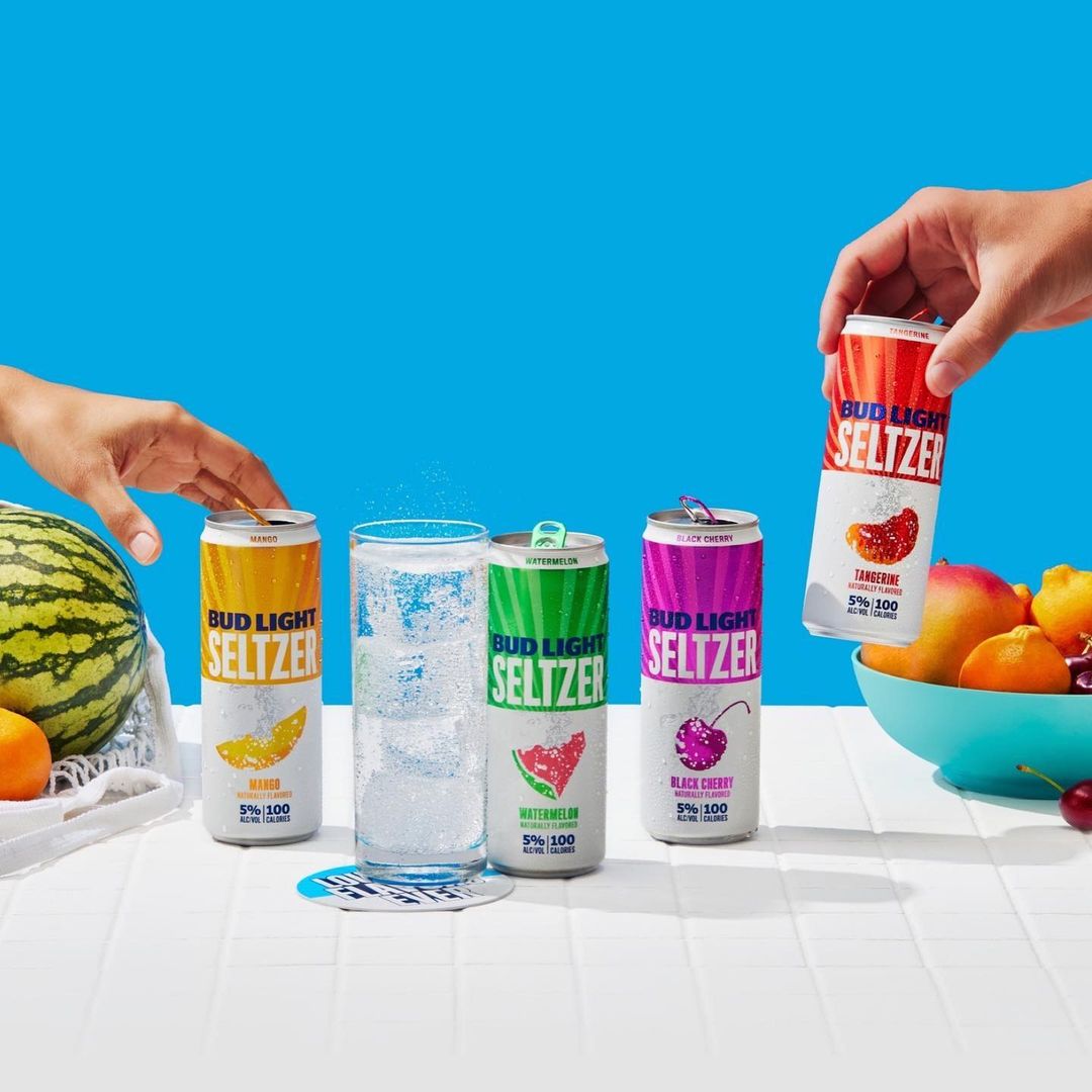Four cans of Bud Light Seltzer are displayed with a full glass and fresh fruit on a white tile surface against a bright blue background.