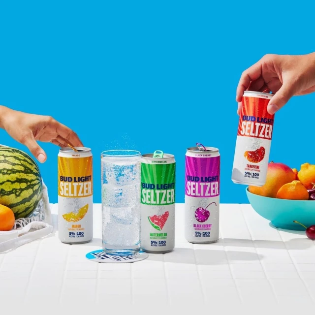 Four cans of Bud Light Seltzer are displayed with a full glass and fresh fruit on a white tile surface against a bright blue background.