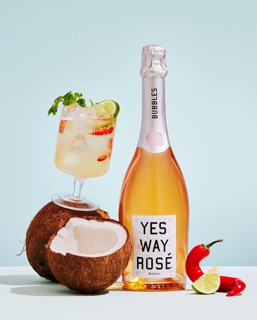 Rosé champagne bottle and cocktail with fresh ingredients, including coconut, lime, mint, and chili peppers, against blue background.