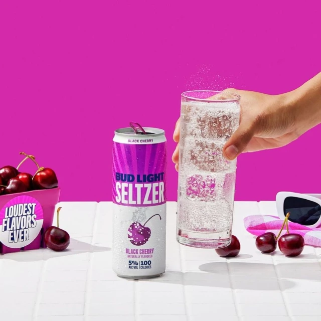 Bud Light Seltzer Black Cherry can and a glass of sparkling liquid are on a white tiled surface against a bright pink background.
