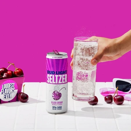 Bud Light Seltzer Black Cherry can and a glass of sparkling liquid are on a white tiled surface against a bright pink background.