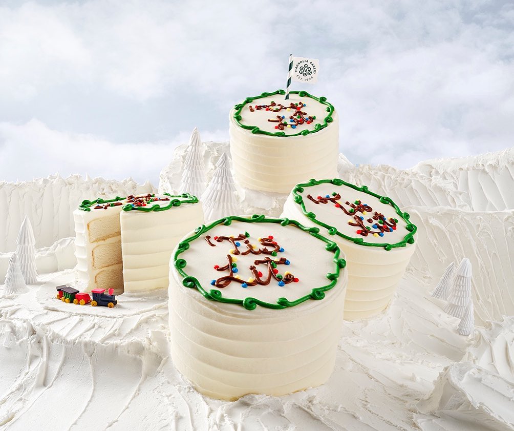 Four tiered cakes with white frosting and colorful sprinkles, set against a snowy landscape with decorative trees and mountain backdrop.