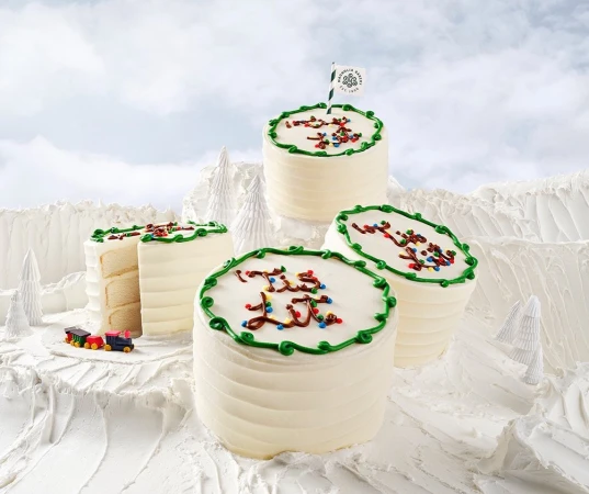 Four tiered cakes with white frosting and colorful sprinkles, set against a snowy landscape with decorative trees and mountain backdrop.