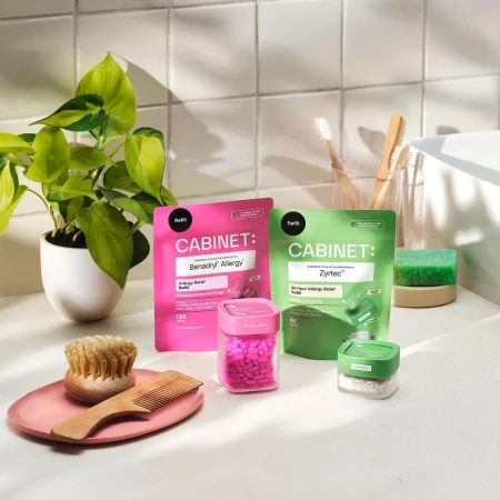 Close-up of pink and green Cabinet allergy relief containers and pills on a white countertop next to a plant in a white pot.