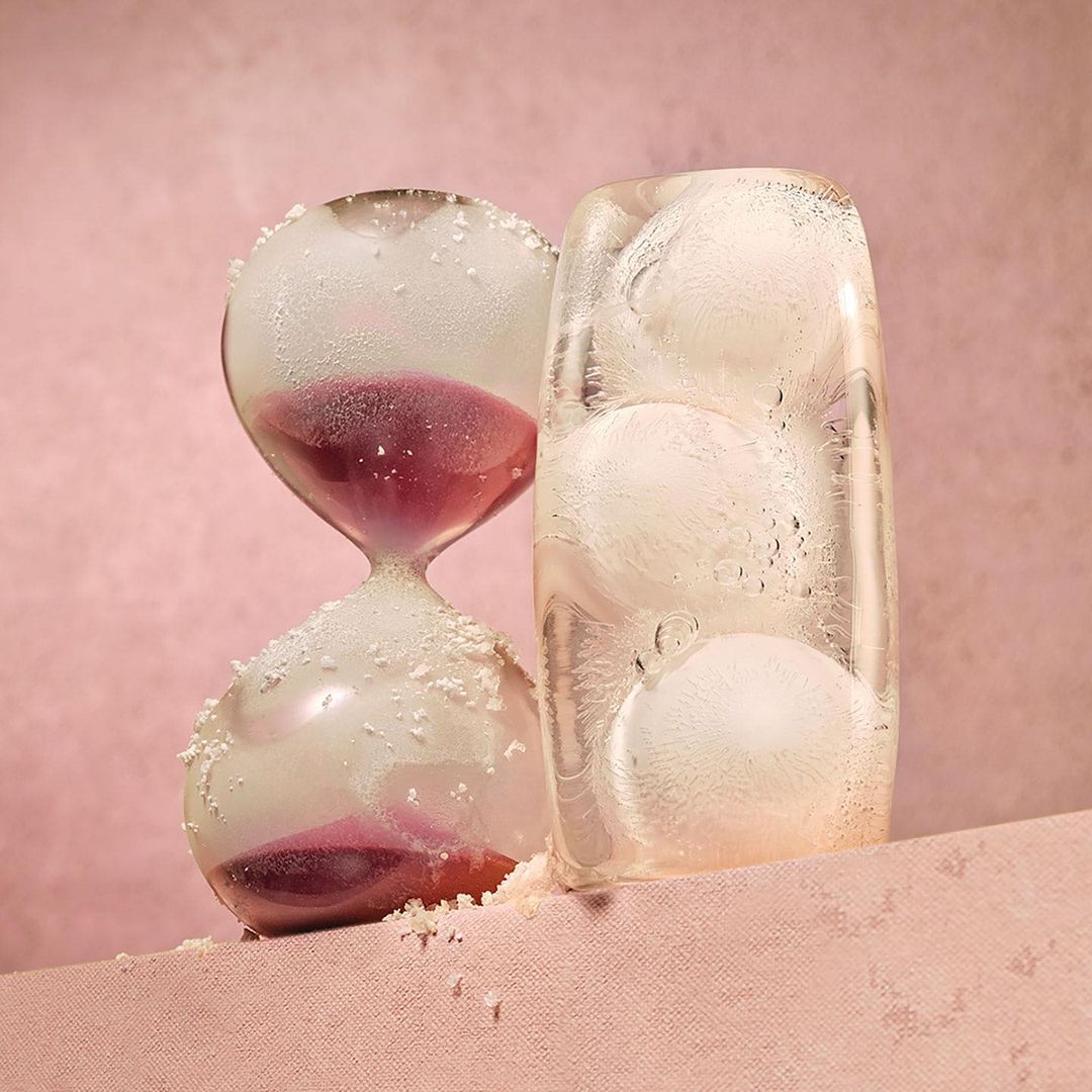 Hourglass with purple sand and ice block with ice balls on pink surface and background in studio lighting casting long shadows.