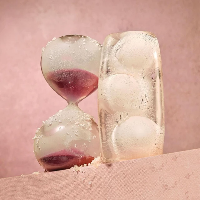 Hourglass with purple sand and ice block with ice balls on pink surface and background in studio lighting casting long shadows.