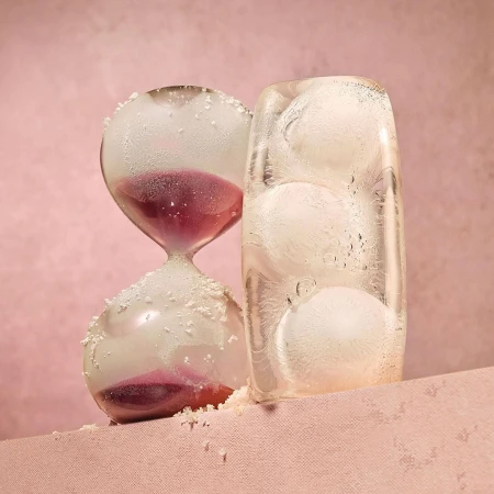 Hourglass with purple sand and ice block with ice balls on pink surface and background in studio lighting casting long shadows.