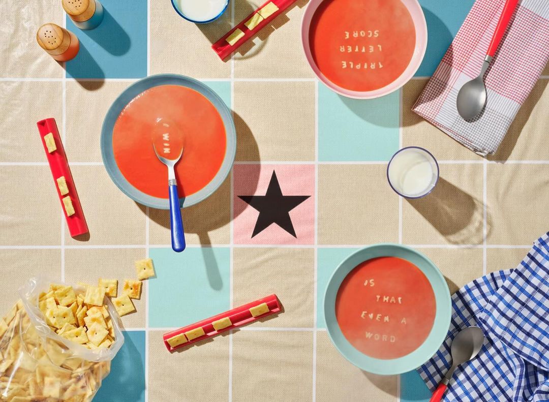 Overhead shot of three bowls of tomato soup with cracker letters spelling words, surrounded by crackers, milk, and dish towels.